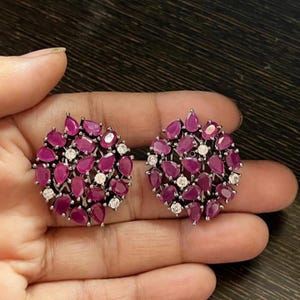 Puede incluir: Un par de pendientes redondos con un racimo de rubíes de color rosa intenso en forma de pera y pequeñas piedras blancas transparentes de corte redondo, engastados en una montura de metal oscuro oxidado con diseño floral.