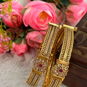 Peut inclure: Deux bracelets rigides dorés présentant un motif en métal tressé et texturé. Chaque bracelet comprend une section rectangulaire centrale ornée d'un motif floral circulaire, serti d'une pierre rouge au centre entourée de petites pierres blanches.