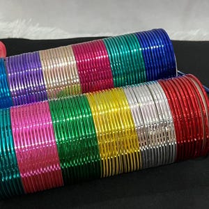 Puede incluir: Dos pilas cilíndricas de brazaletes de metal delgados en una variedad de colores vibrantes que incluyen azul, morado, dorado, rosa, verde, plateado y rojo. Los brazaletes tienen un acabado liso y reflectante y están dispuestos en filas.