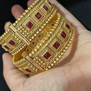 May include: A pair of gold-toned hinged bangles featuring a repeating pattern of square-cut deep red stones set within textured metal frames. Each bracelet displays a series of raised gold beads and intricate linear engravings along the edges.