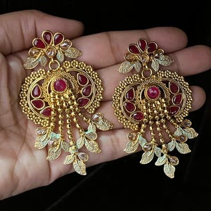 Könnte beinhalten: Ein Paar goldfarbene Ohrhänger mit einem blumenartigen Oberteil und roten tropfenförmigen Steinen. Das runde Hauptteil besteht aus filigranen Metallarbeiten, zentralen roten Steinen und herabhängenden Ketten mit blattförmigen Anhängern.
