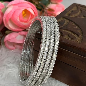 Op de afbeelding: Een set van vier zilverkleurige armbanden, elk voorzien van een doorlopende rij kleine, heldere, rondgeslepen stenen die in de metalen band zijn gezet. De armbanden zijn cirkelvormig en hebben een reflecterende, metalen afwerking.
