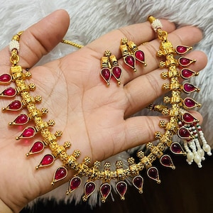 Könnte beinhalten: Ein goldfarbenes Halsketten- und Ohrring-Set. Die Halskette und die Ohrringe sind mit tropfenförmigen, tiefroten Edelsteinen versehen. Der Schmuck wird auf einer Hand präsentiert, die das komplizierte Design und die Handwerkskunst zeigt. Das Set enthält eine Halskette und ein Paar Ohrringe.