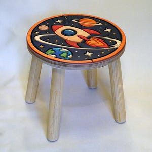 Pode incluir: Um banco redondo de madeira com quatro pernas de cor clara. O assento apresenta um design colorido incrustado de uma cena espacial com um foguete, planetas e estrelas sobre um fundo azul escuro, emoldurado por uma borda circular laranja.