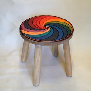 May include: A small wooden stool with four cylindrical legs and a round seat featuring a vibrant, multi-coloured spiral pattern. The spiral design transitions through shades of red, orange, yellow, green, blue, and purple, creating a swirling effect.