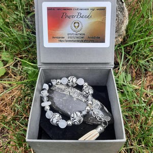 Könnte beinhalten: Ein Perlenarmband mit schwarz-weiß gemusterten Perlen, durchscheinenden weißen Perlen und silberfarbenen Akzenten, ausgestattet mit einem kleinen silbernen Blattanhänger und einer beigen Quaste, präsentiert in einer grauen Box mit PowerBands-Etikett.