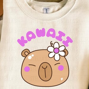 May include: Cream-colored sweatshirt with a Kawaii capybara graphic. The design features a brown capybara face with a flower, pink cheeks, and the word "KAWAII" in purple. The Japanese word "カピバラ" is below the capybara.