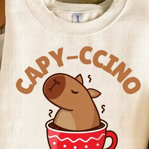 May include: Cream-colored sweatshirt with a playful graphic. The design features a cartoon capybara in a red coffee cup, with the word "CAPY-CCINO" in brown letters above. A fun, cozy top for animal lovers.