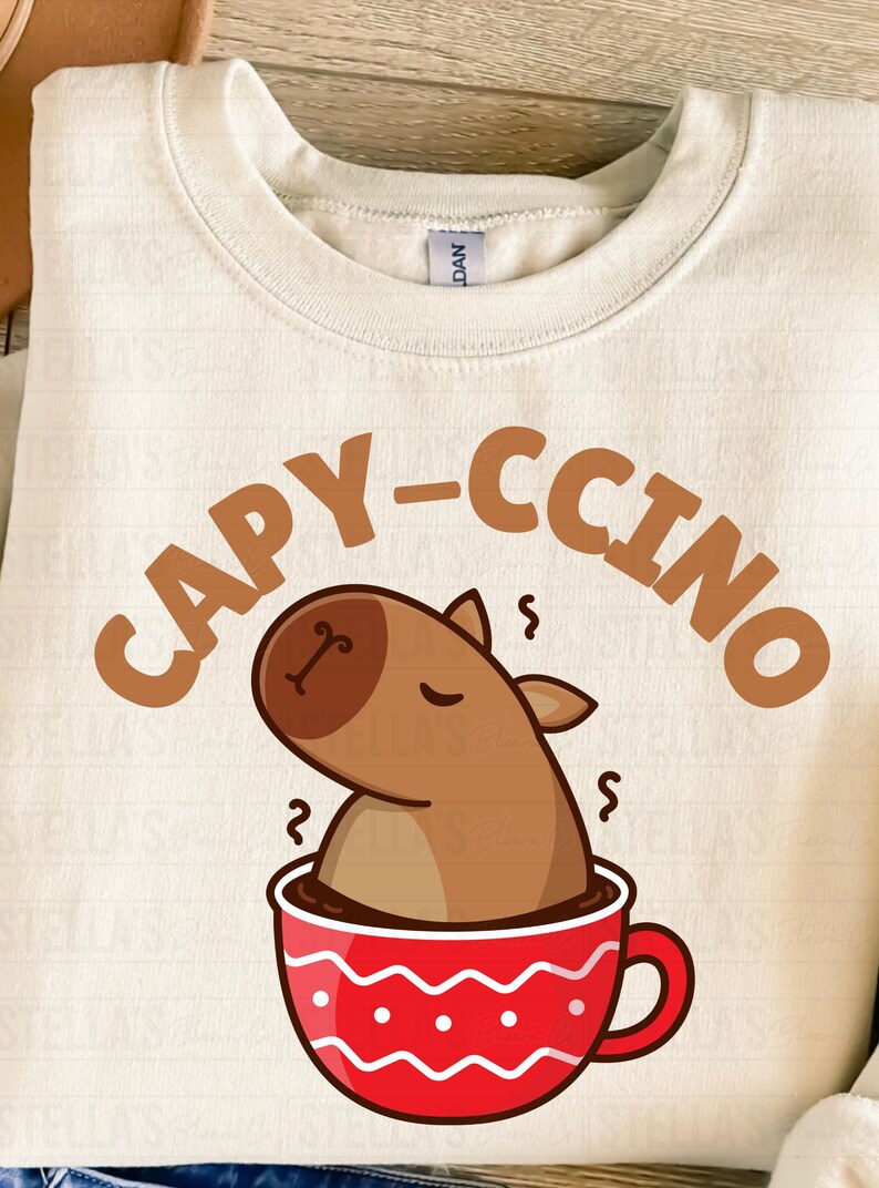 May include: Cream-colored sweatshirt with a playful graphic. The design features a cartoon capybara in a red coffee cup, with the word "CAPY-CCINO" in brown letters above. A fun, cozy top for animal lovers.