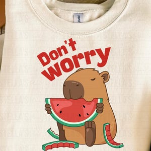 May include: Cream-colored sweatshirt with a graphic of a capybara eating watermelon. The text "Don't Worry, Be Capy" is printed in red. The capybara is brown with a red and green watermelon slice.