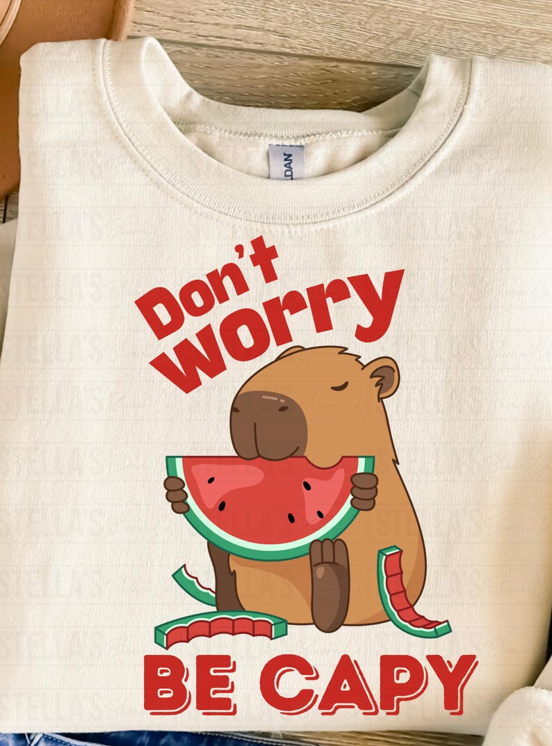 May include: Cream-colored sweatshirt with a graphic of a capybara eating watermelon. The text "Don't Worry, Be Capy" is printed in red. The capybara is brown with a red and green watermelon slice.