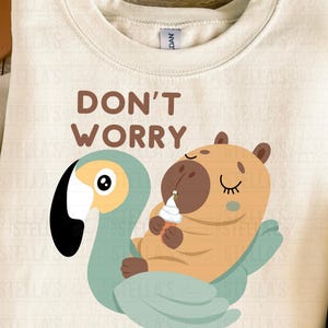 May include: Cream-colored sweatshirt with a whimsical illustration. The design features a cartoon capybara holding an ice cream cone, riding a teal swan. Text reads "DON'T WORRY BE CAPPY" in brown lettering.