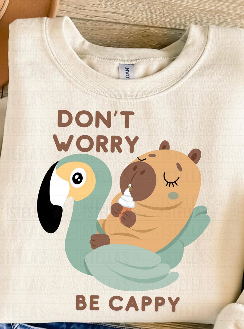 May include: Cream-colored sweatshirt with a whimsical illustration. The design features a cartoon capybara holding an ice cream cone, riding a teal swan. Text reads "DON'T WORRY BE CAPPY" in brown lettering.