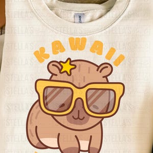 May include: Cream-colored sweatshirt featuring a cartoon capybara wearing yellow sunglasses and a star. The word "KAWAII" is above the capybara, and Japanese text is below. A fun, whimsical design.