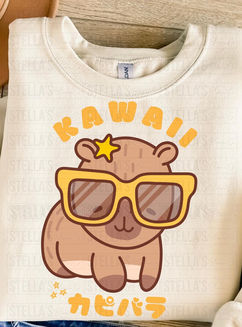 May include: Cream-colored sweatshirt featuring a cartoon capybara wearing yellow sunglasses and a star. The word "KAWAII" is above the capybara, and Japanese text is below. A fun, whimsical design.