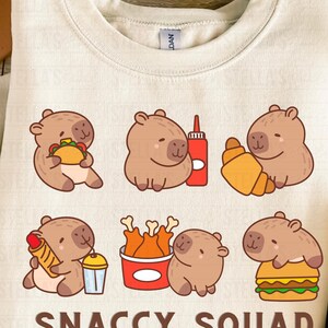 May include: Cream-colored sweatshirt featuring a playful graphic of cartoon capybaras with various foods like tacos, croissants, and burgers. The text "SNACCY SQUAD" is printed at the bottom in a bold font. A fun, food-themed design.