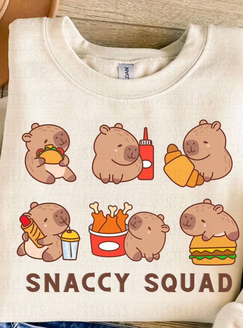 May include: Cream-colored sweatshirt featuring a playful graphic of cartoon capybaras with various foods like tacos, croissants, and burgers. The text "SNACCY SQUAD" is printed at the bottom in a bold font. A fun, food-themed design.