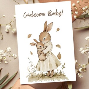 May include: A greeting card with the text "Welcome Baby!" features a watercolor illustration of two brown bunnies. One larger bunny holds a smaller one, surrounded by leaves and grass, in a soft, neutral color palette.