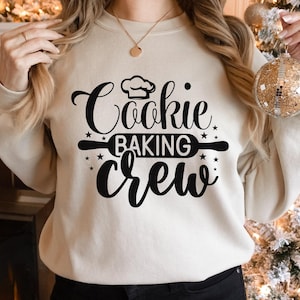May include: A cream-colored sweatshirt with the words "Cookie Baking Crew" in black script and a chef's hat graphic. The design includes a rolling pin and star accents, perfect for baking enthusiasts.