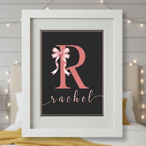 May include: A framed print featuring a large, coral-colored letter "R" adorned with a pink bow on a black background. The name "Rachel" is written in cursive below the letter. The print is in a white frame.