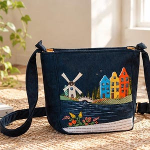 May include: A dark blue denim crossbody bag featuring an embroidered scene of a white windmill, a small bridge, and three colourful houses in red, yellow, and blue. The bottom of the bag has a white patterned border with floral embroidery.