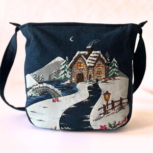 以下が含まれることがあります： 冬の風景が刺繍されたダークブルーの布製ショルダーバッグ。石造りのコテージ、明かりの灯る窓、雪に覆われた木々、暗い川に架かる石橋、三日月の下にある街灯が描かれています。