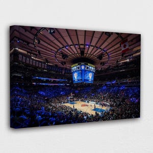 May include: A canvas print of a basketball arena filled with spectators. The arena has a large scoreboard and a circular ceiling. The crowd is illuminated with blue lights, and the court is visible in the center.