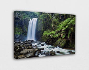 May include: A canvas print of a waterfall cascading into a rocky stream. Lush green ferns and trees surround the falls, creating a serene natural landscape. The water appears smooth and flowing, with a soft, blurred effect.
