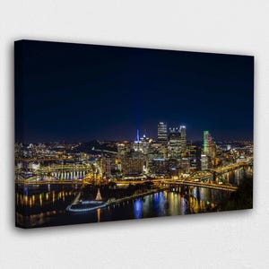 May include: A canvas print of a nighttime cityscape. The image showcases a vibrant city skyline with illuminated buildings, bridges, and reflections in the water. The dark blue sky contrasts with the bright city lights.
