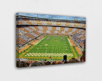 May include: A canvas print of a packed football stadium with orange and white seating. The green field has yard lines and a checkerboard end zone. A marching band is on the field. The sky is blue with clouds.