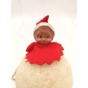 May include: A vintage Christmas ornament featuring a doll-like figure. The doll has a painted face with blue eyes, wearing a red and white Santa hat and a red felt cape. The body is a cream-colored, rounded shape, possibly made of foam.