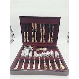 May include: A flatware set featuring silver-toned heads and gold-toned handles with a bamboo-inspired texture. The set includes knives, forks, and spoons, organized within a dark wooden storage case lined with deep burgundy velvet fabric.