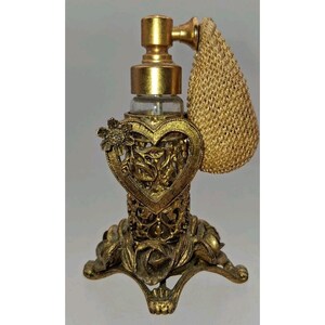 May include: A gold-toned perfume atomizer featuring a clear glass bottle encased in an ornate metal frame with a heart-shaped cutout, a floral accent, and a textured base. A beige mesh bulb is attached to the top of the metallic spray nozzle.