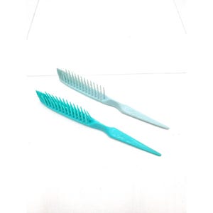 May include: Two plastic hair combs. One is a light blue color, and the other is a bright teal. Both combs have a long handle and a wide head with many teeth. The combs are angled and appear to be new.