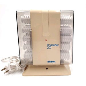 May include: A vintage beige hairsetter with the text "Hairsetter 20" and the brand name "belson." The hairsetter has clear plastic compartments holding white hair rollers. A white power cord with a two-prong plug is attached.