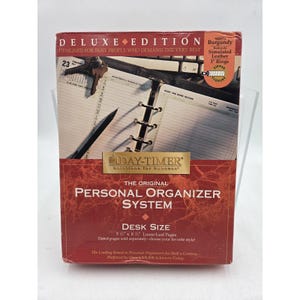 May include: A burgundy Day-Timer personal organizer system in a deluxe edition box. The packaging features a graphic of an open planner with 1-inch rings, a pen, and keys. It specifies desk size for 5 1/2 inch by 8 1/2 inch loose-leaf pages.