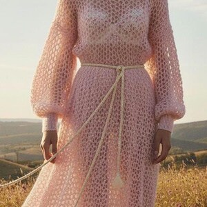 May include: A light pink, open-weave dress with long sleeves and a floor-length skirt. The dress is cinched at the waist with a cream-colored rope belt. The model is standing in a field of tall grass and wildflowers.