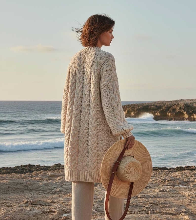 May include: A cream-colored, cable-knit cardigan with long sleeves and a knee-length design. A straw sun hat with a brown leather strap is held in the hand. The background features a beach and ocean.