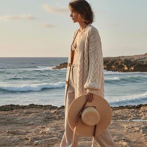 May include: A woman walking on a beach, wearing a cream-colored cable-knit cardigan, beige pants, and holding a straw hat with a brown leather strap. The ocean and rocky coastline are in the background.