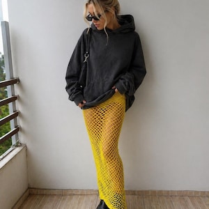 May include: A person wearing a dark gray hoodie, black sunglasses, and a yellow fishnet skirt. The person is also wearing black platform boots. The outfit is styled in a casual, yet fashionable manner.