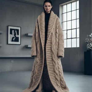 Könnte beinhalten: Langer, beigefarbener Zopfstrick-Cardigan mit Schalkragen. Der bodenlange Pullover hat ein dickes, strukturiertes Gewebe und lange Ärmel. Das Model trägt ein schwarzes Oberteil und Stiefel. Der Hintergrund ist ein grauer Raum mit Fenster und Kunstwerk.