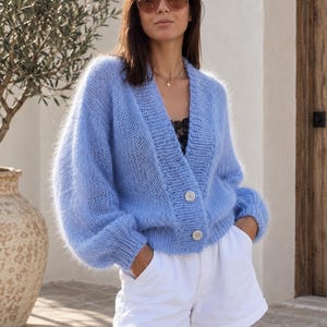 Peut inclure: Un cardigan bleu clair, duveteux, à manches longues, avec une encolure en V et deux gros boutons sur le devant. Le vêtement en tricot présente des poignets et un ourlet côtelés, associé à un short blanc uni et un haut en dentelle noire.