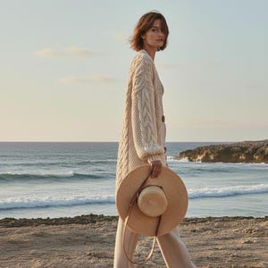 May include: A woman walking on a beach, wearing a cream-colored cable-knit cardigan and beige pants. She carries a wide-brimmed straw hat with a brown leather strap. The background features the ocean and a clear sky.