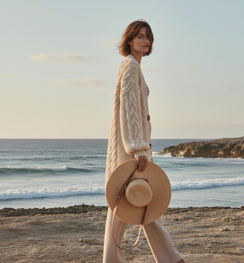 May include: A woman walking on a beach, wearing a cream-colored cable-knit cardigan and beige pants. She carries a wide-brimmed straw hat with a brown leather strap. The background features the ocean and a clear sky.