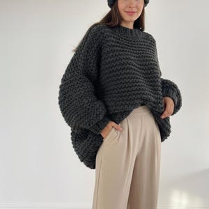 Puede incluir: Una mujer con un suéter de punto grueso gris oscuro y un gorro a juego. El suéter tiene un ajuste amplio y de gran tamaño con mangas largas. Lleva pantalones anchos de color beige claro. El atuendo es de estilo casual y acogedor.