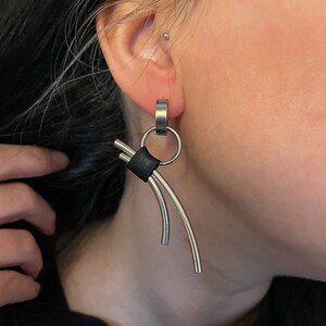 Peut inclure: Boucles d'oreilles créoles argentées avec un accent en cuir noir et deux barres argentées courbées suspendues à la créole.