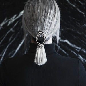 May include: A person with grey hair wearing a black turtleneck sweater and a silver triangle hair clip.
