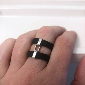 May include: A black ring with a silver band and a rectangular cutout. The ring is worn on a person's finger.