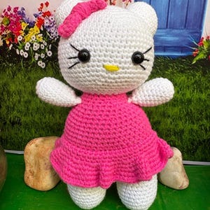 May include: A handmade white crochet doll with a pink dress and bow. The doll has black eyes, a yellow beak, and black whiskers. The doll is standing on a green surface with rocks in the background. The doll is a cute, handmade item.