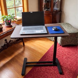 May include: A brown laptop desk with a black base and a dark wood-grain surface. A laptop, blue mousepad, and mouse are on the desk. The desk is in front of a window with a plant.
